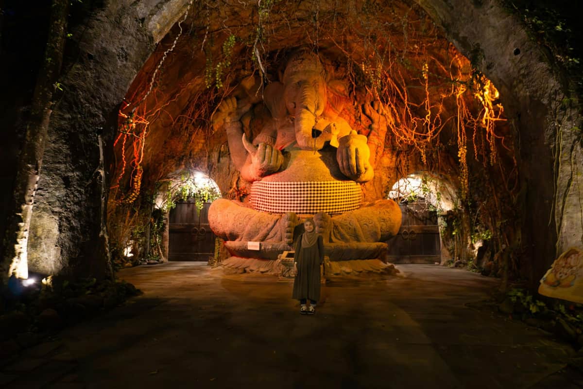 A person standing in front of a large, intricately carved statue of Ganesha in a dimly lit, cavern-like setting.