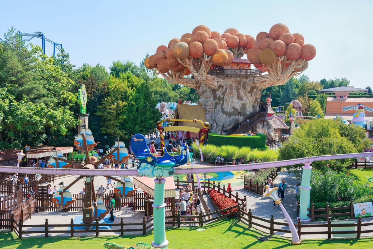 Amusement park with colorful rides, large tree structure, and people enjoying various attractions under a clear blue sky.