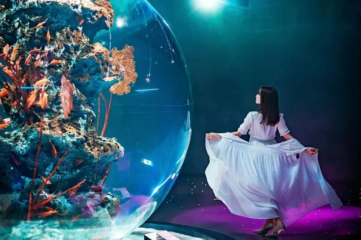 átoa: Entry Ticket A woman in a white dress stands near a large illuminated glass sphere containing coral, in a dimly lit environment.