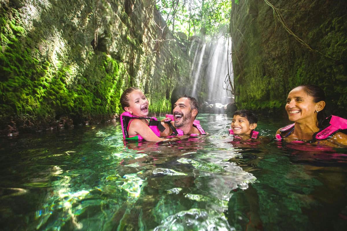A family of four wearing life jackets smiles and enjoys swimming in a green canyon with sunlight streaming through trees above.