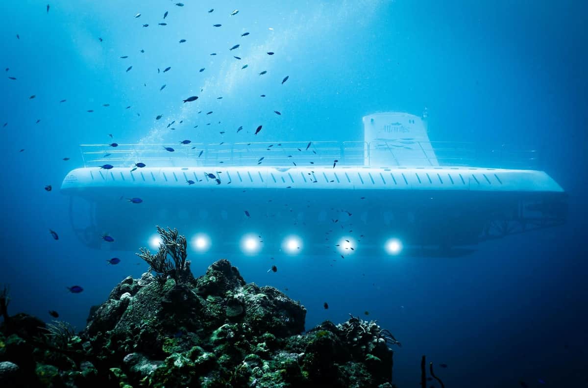 Atlantis Cozumel: Submarine Experience A submerged white submarine with illuminated windows glides past rocky coral, surrounded by small fish in clear blue water.