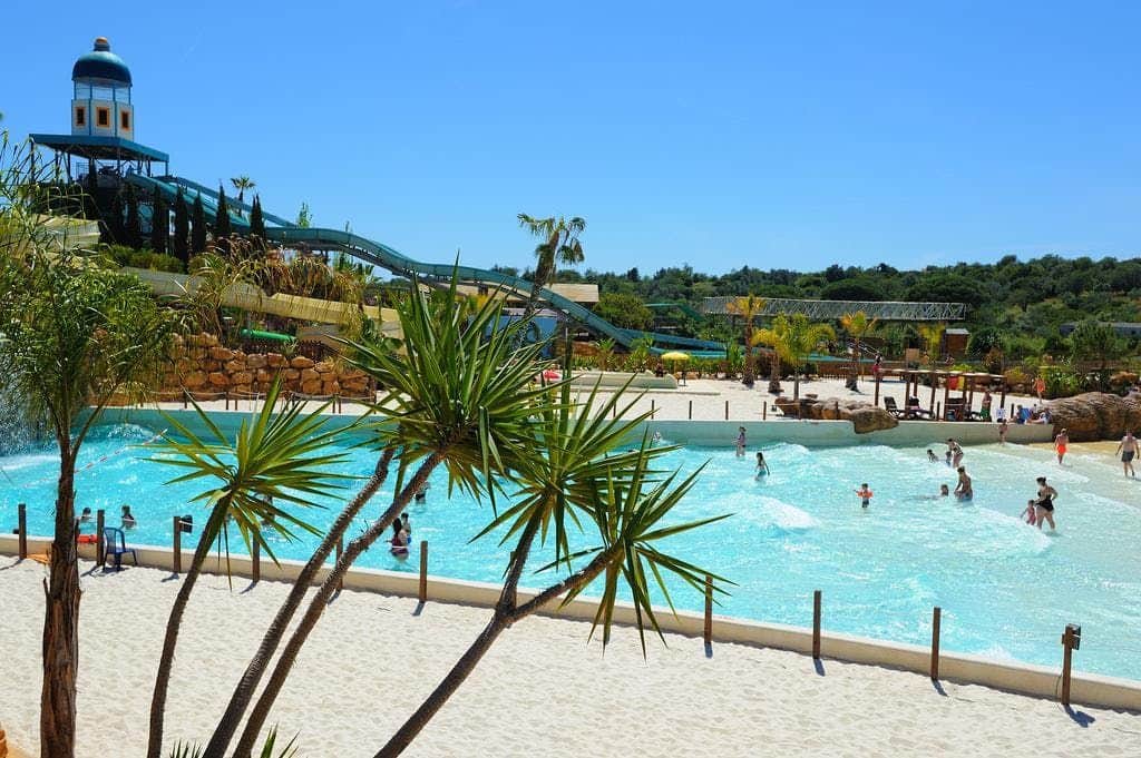 A water park with people swimming in a wave pool, surrounded by palm trees and water slides under a clear blue sky.