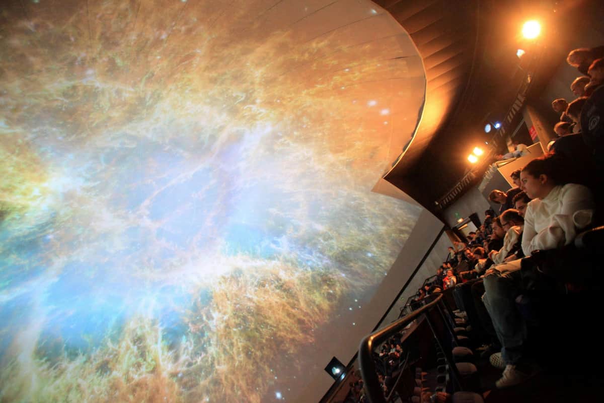 People sitting in a planetarium, watching a projection of a cosmic nebula on a large domed ceiling.