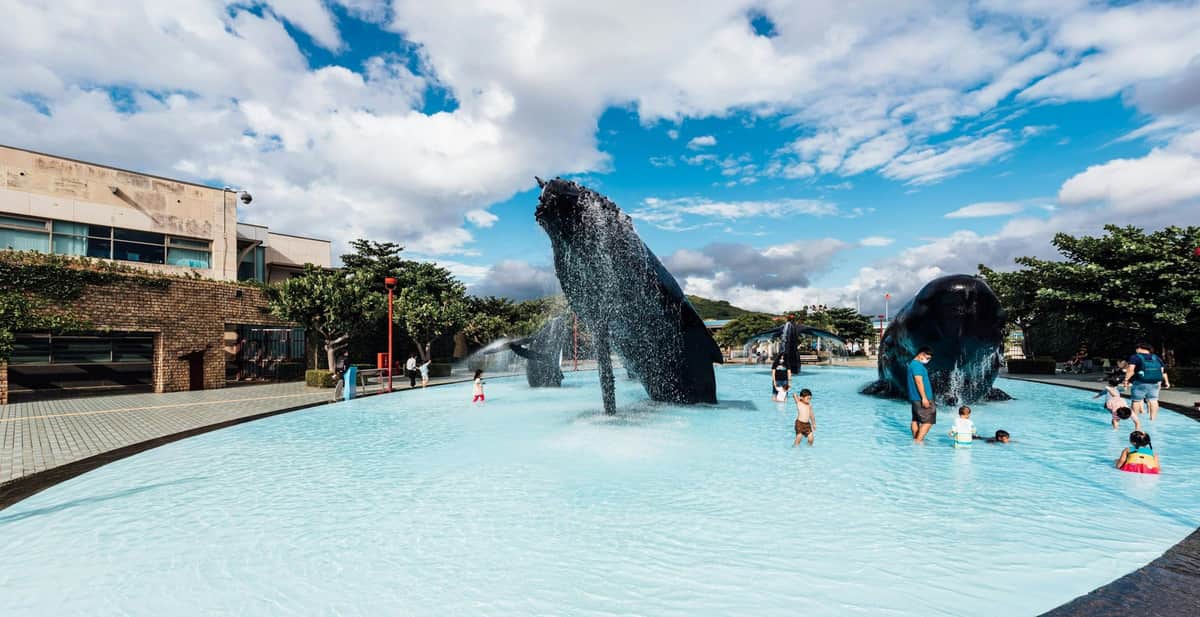 A fountain featuring a whale sculpture spouting water in a shallow pool with people enjoying the surroundings under a blue sky.