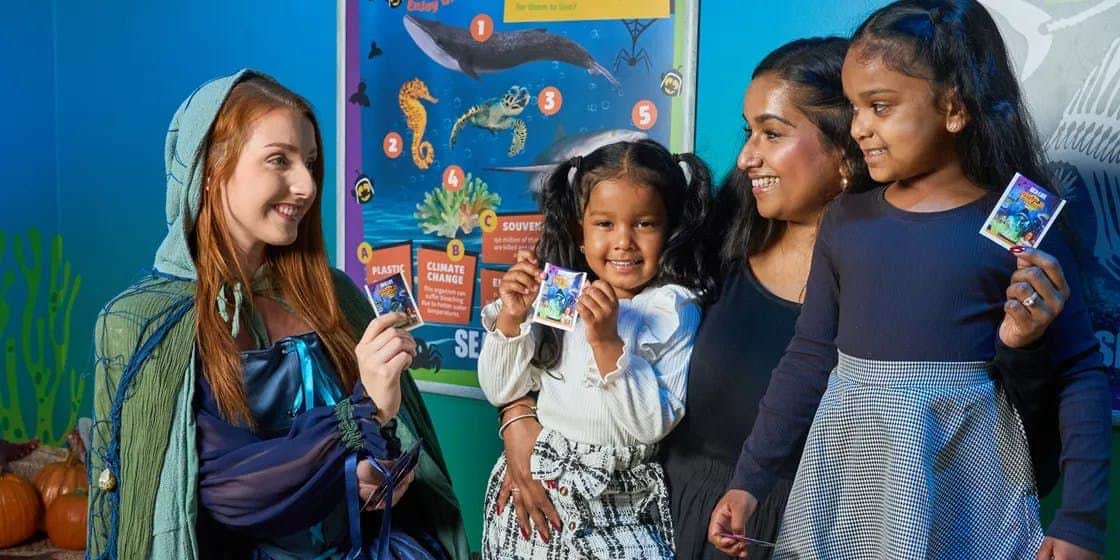 SEA LIFE Michigan: Entry Ticket Three children and a woman smiling near a colorful educational poster about marine life. One child holds up a card.