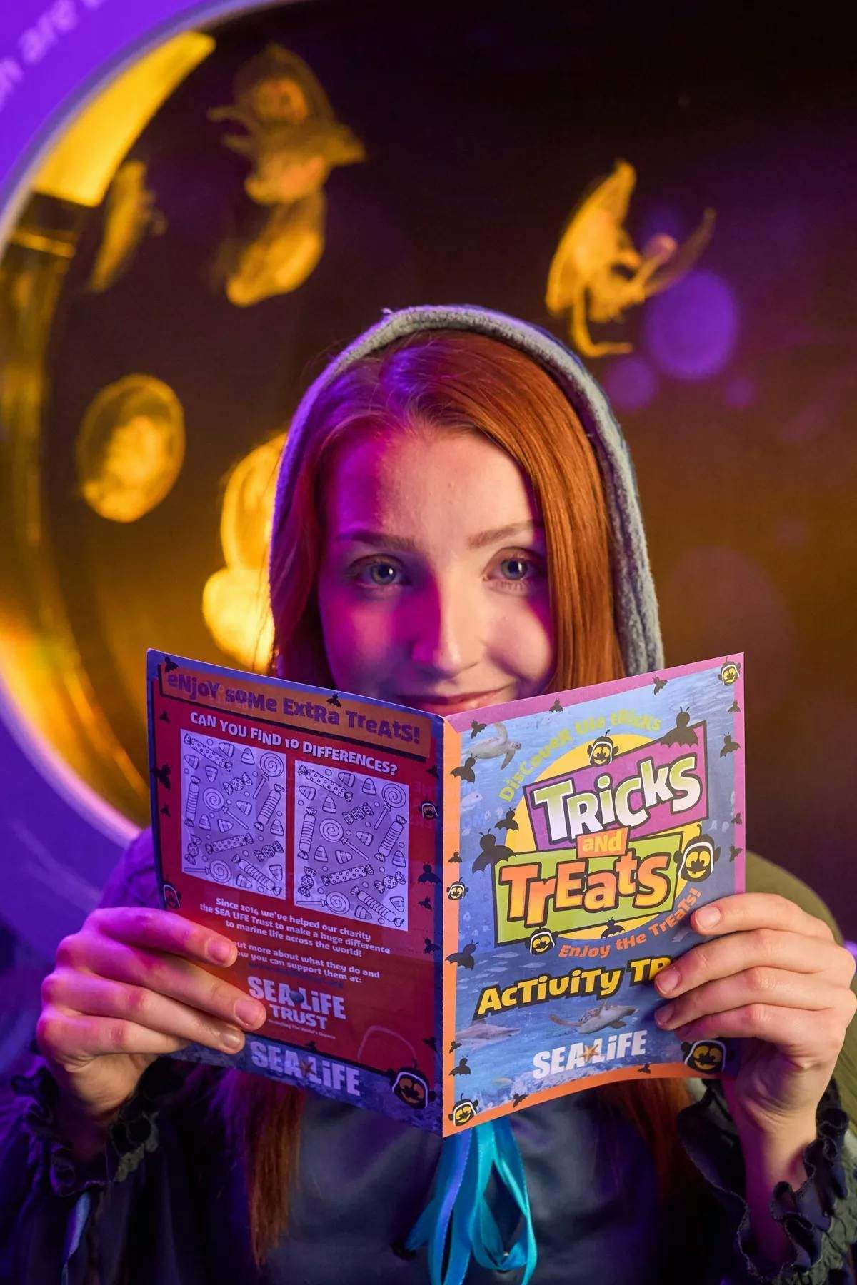SEA LIFE Grapevine A person in a hoodie holds a colorful activity booklet titled "Tricks and Treats" with an aquarium backdrop.