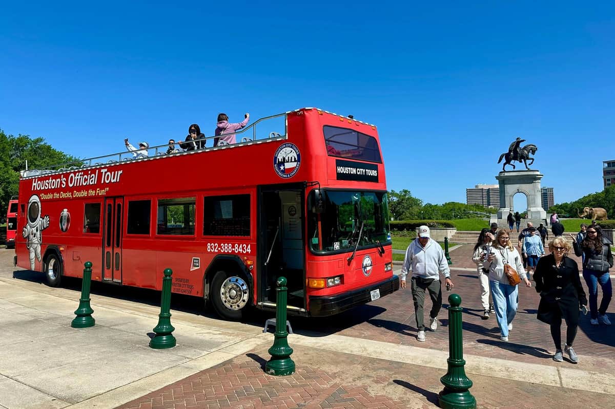 Houston: City Bus Tour + Downtown Aquarium Entry Ticket A red double-decker tour bus parked near a statue of a rider on horseback, with people walking around under a clear blue sky.
