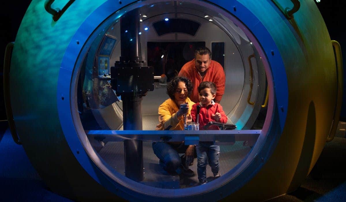 New York Aquarium: Entry Ticket Three people, two adults and a child, interact inside a large, lit, circular structure with control panels and buttons.