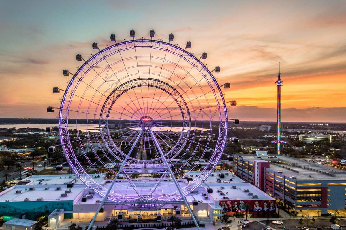 A large ferris wheel lit with purple lights at sunset, with a cityscape and a tall illuminated tower in the background.