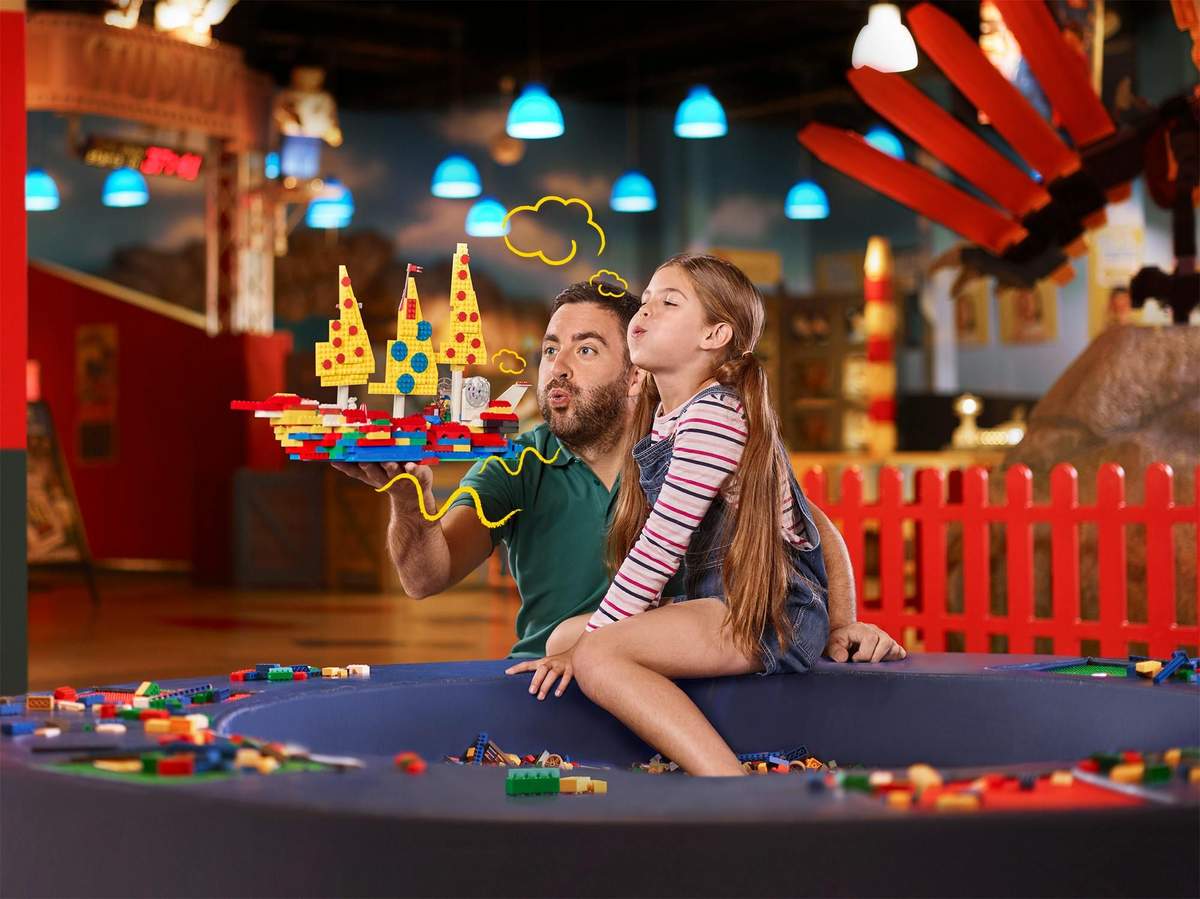 Man and young girl in a LEGO play area, creating a structure together, surrounded by colorful bricks, with drawings of puffs and lines.
