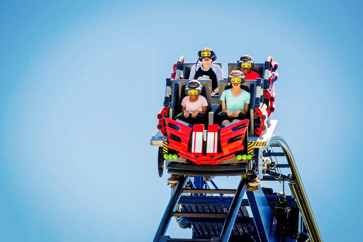 Four people wear VR headsets while riding a colorful, futuristic rollercoaster under a clear blue sky.