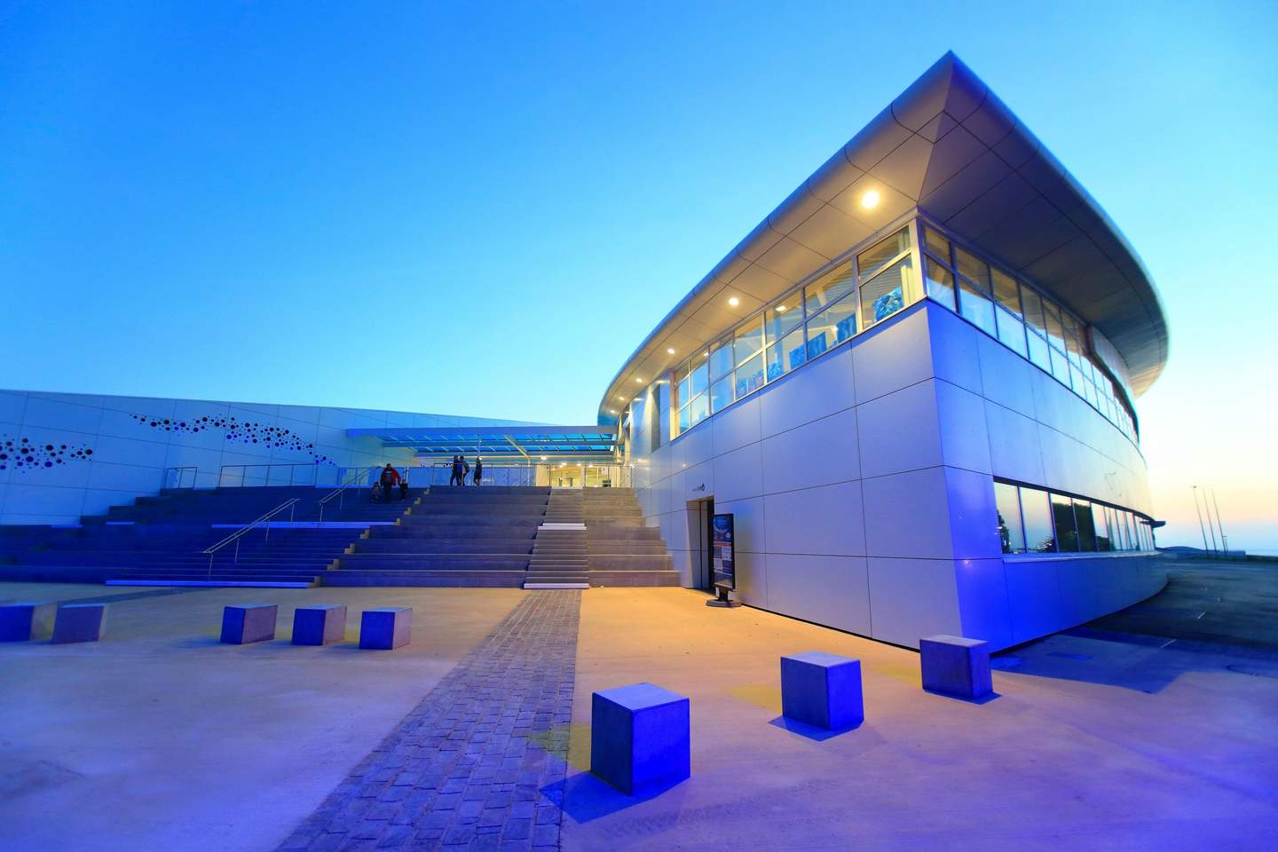 Nausicaá Modern building with large glass windows, concrete steps, and a paved walkway at dusk. Few people are visible on the stairs.