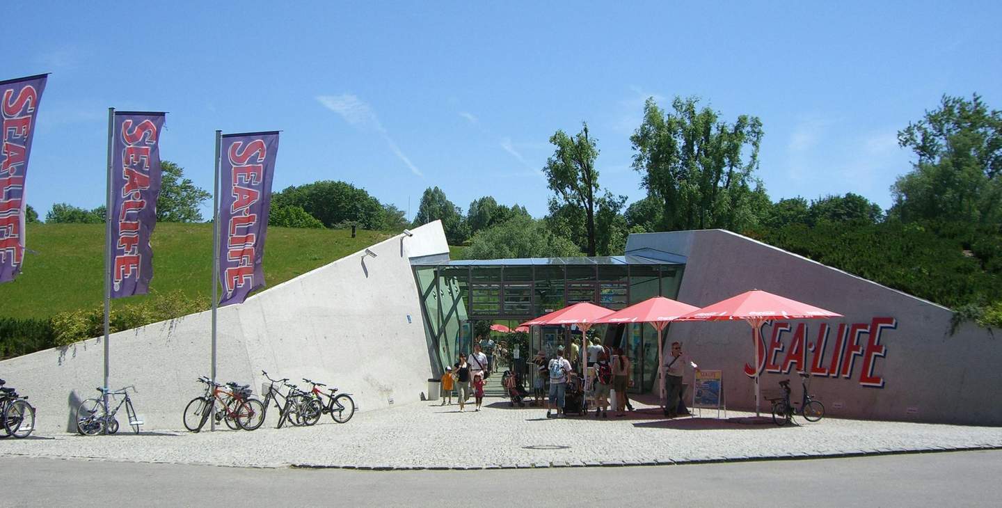 SEA LIFE Munich People gather under red umbrellas outside a building marked "SEA LIFE" with bikes parked nearby. Greenery and flags are visible.