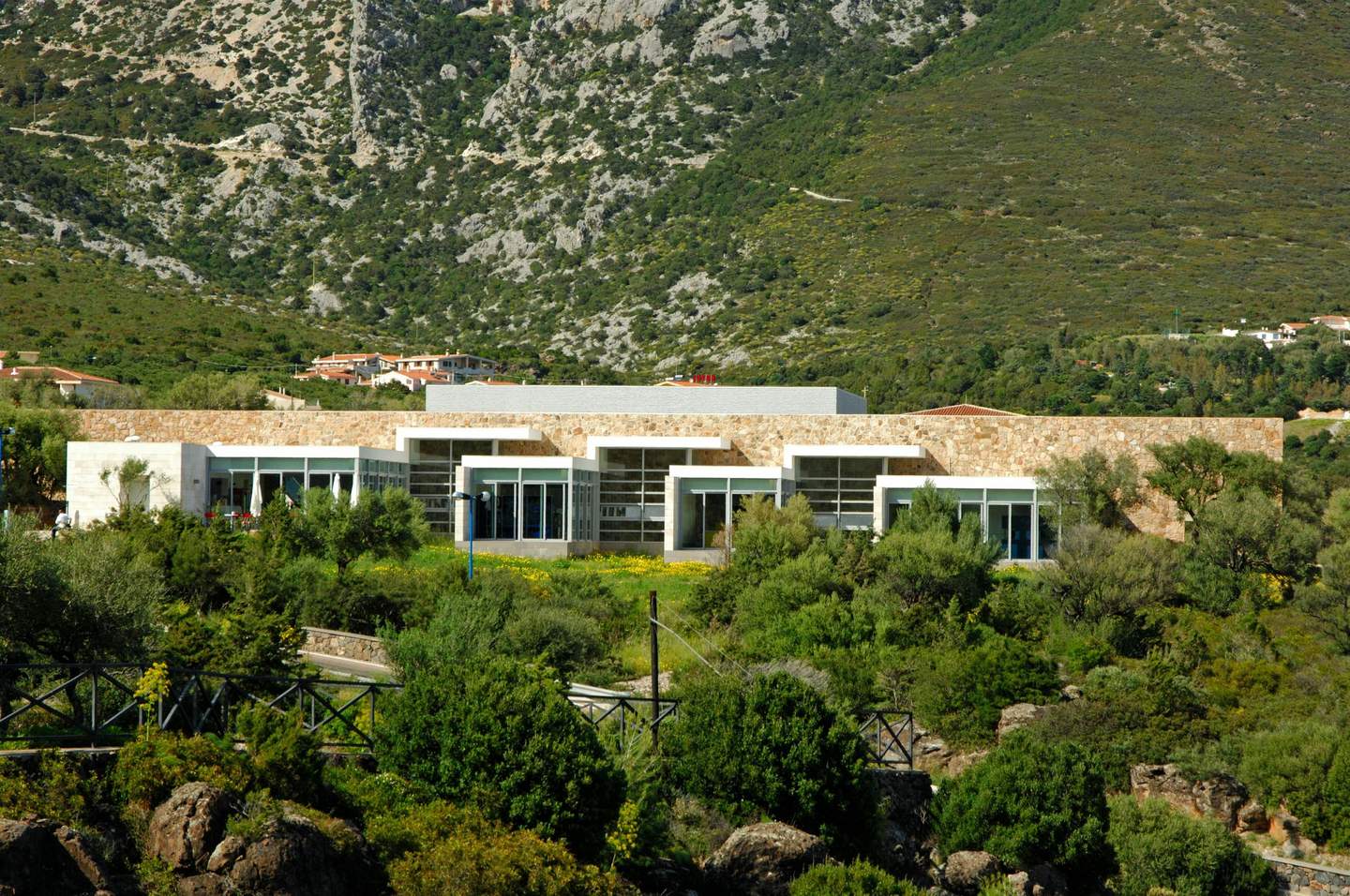Aquarium of Cala Gonone A modern building with large windows and stone facade is set against a mountainous, green landscape with scattered buildings.