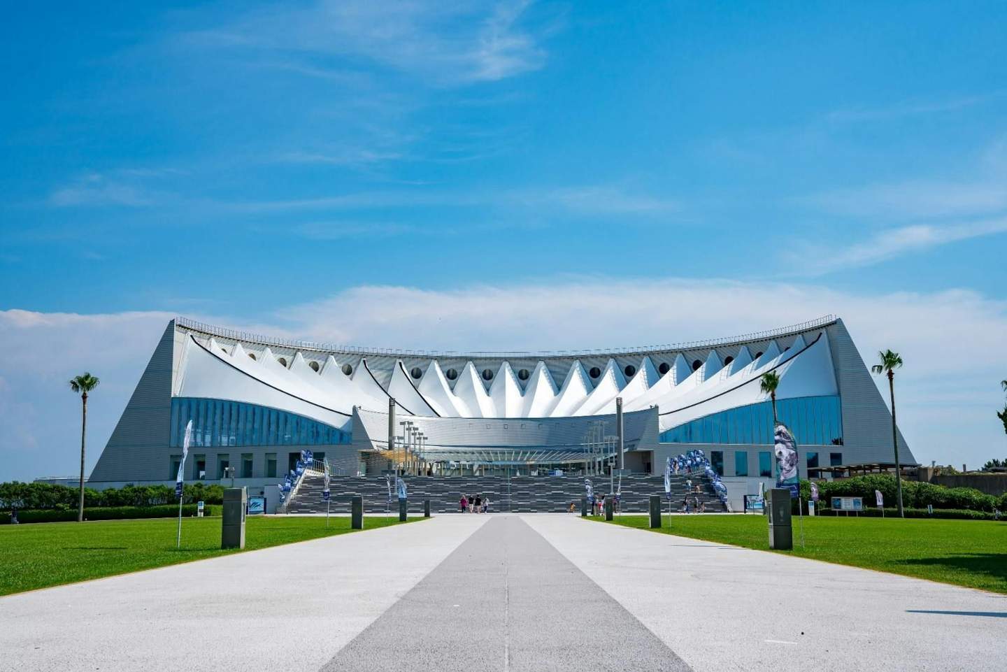 A modern stadium with a white roof and triangular design, seen from a walkway lined with grass and flags under a blue sky.