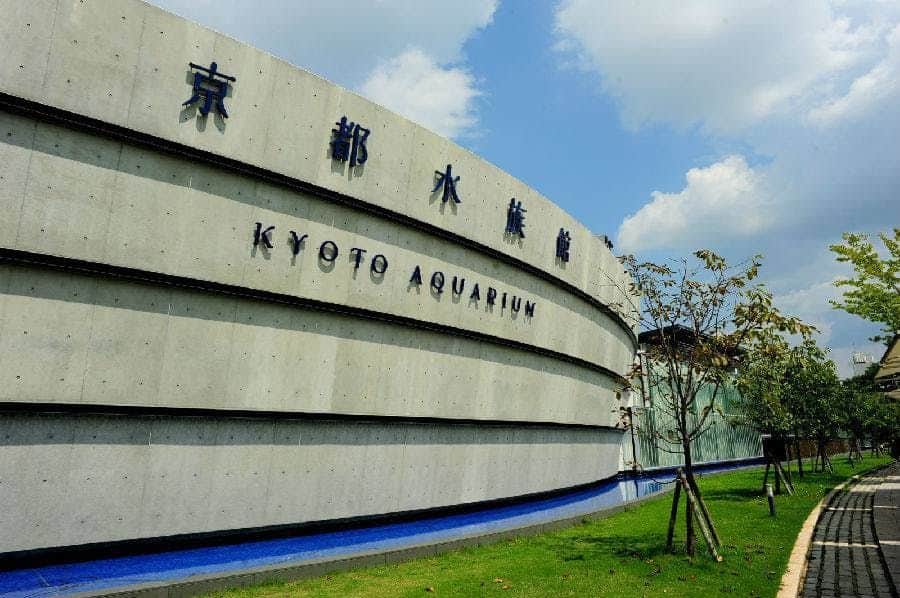 Curved concrete facade of Kyoto Aquarium with trees and a pathway in front, under a partly cloudy sky.