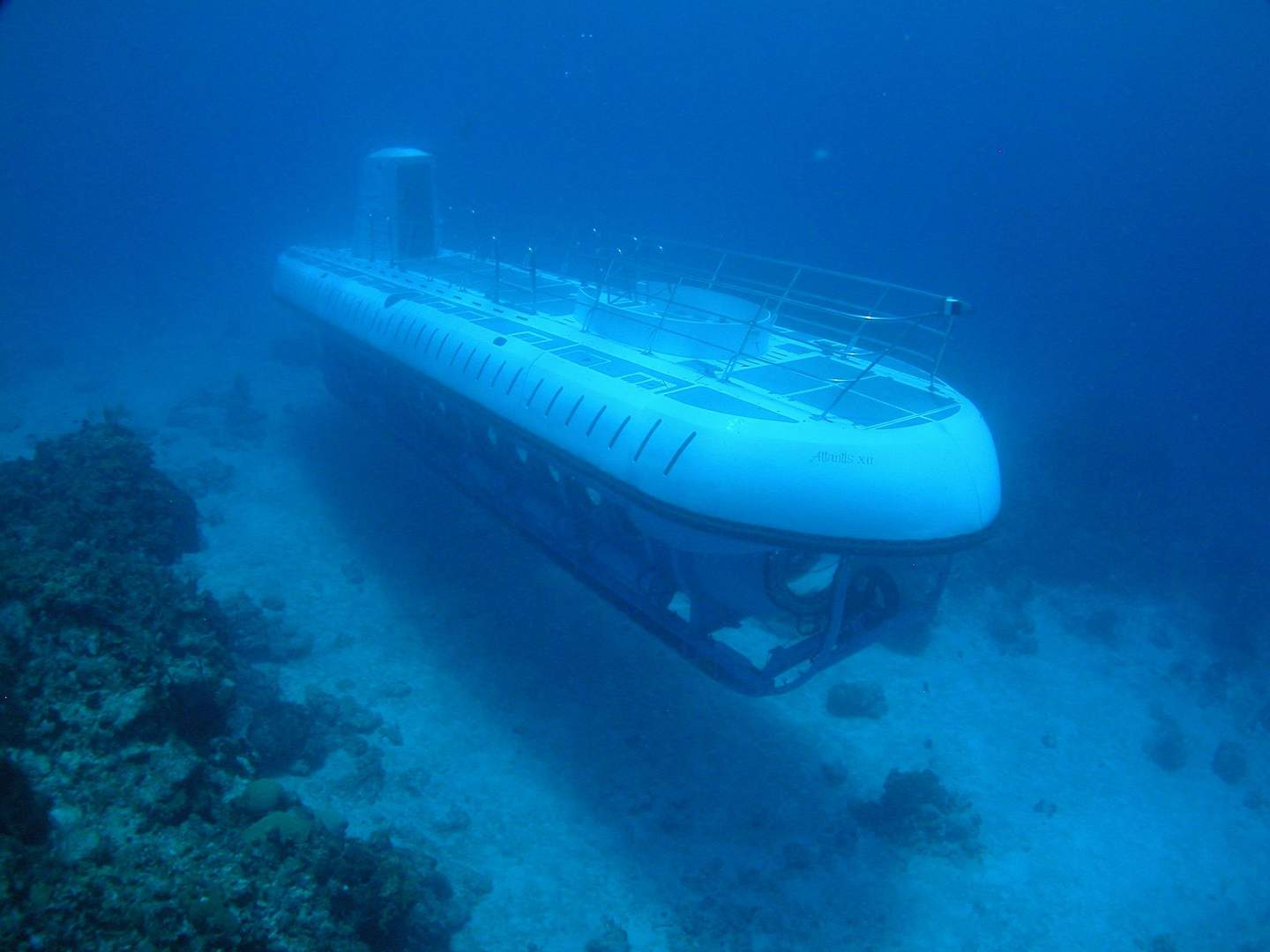 Atlantis Submarines A white submarine underwater, viewed from the side, navigating above the ocean floor with scattered rocks and coral.