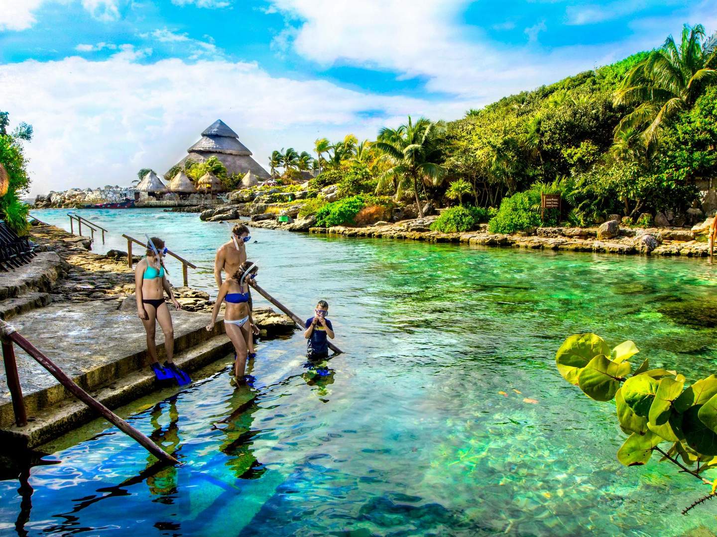 Xel-Há Park People in swimwear and snorkels preparing to enter a clear, tropical lagoon surrounded by lush greenery and thatched-roof structures.