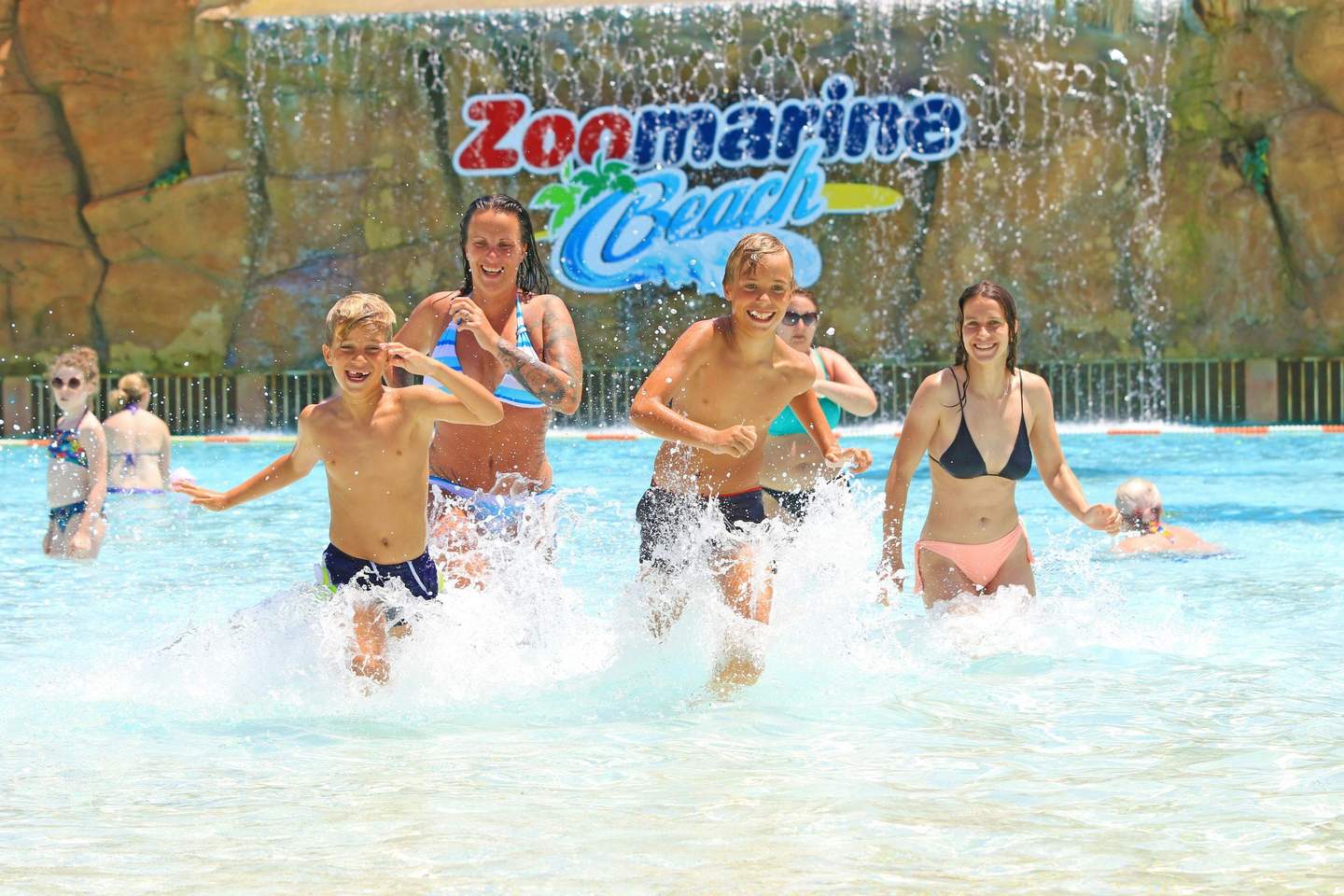 People splashing and playing in a water park area with a "Zoomarine Beach" sign and artificial waterfall in the background.