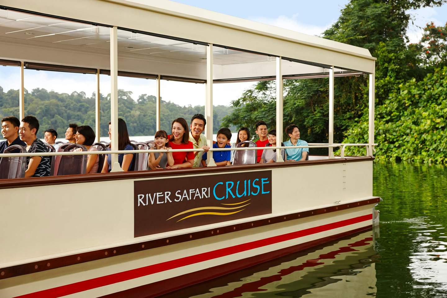 Persone sedute su una barca per il River Safari Cruise, circondata da una vegetazione lussureggiante e da acque calme. Il cartello recita "River Safari Cruise".