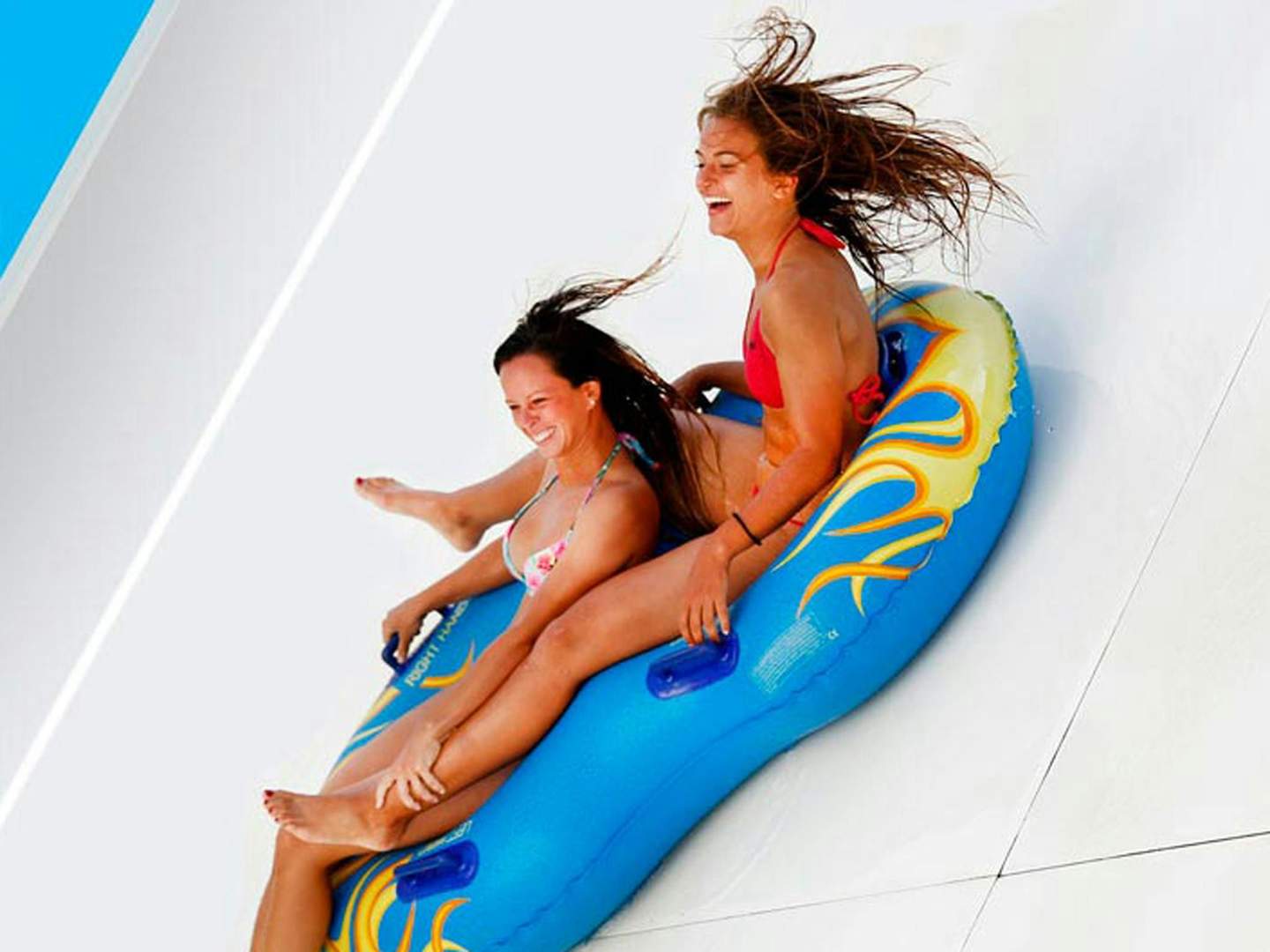 Marineland Catalunya Two women in swimsuits laughing as they slide down a water slide on an inflatable tube.