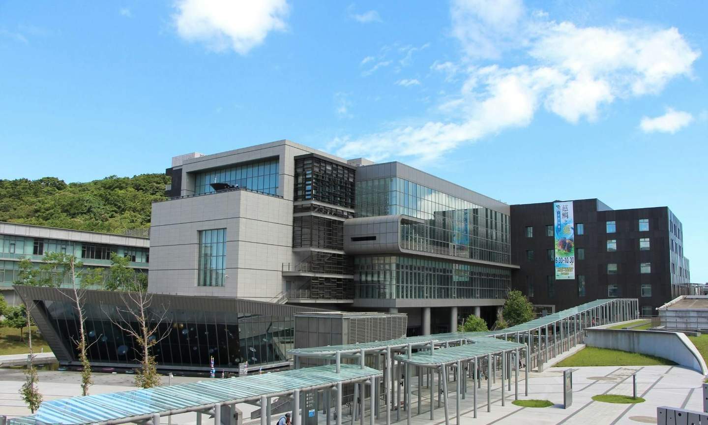 Modern building complex with large glass facades under a clear blue sky, surrounded by greenery and walkways.