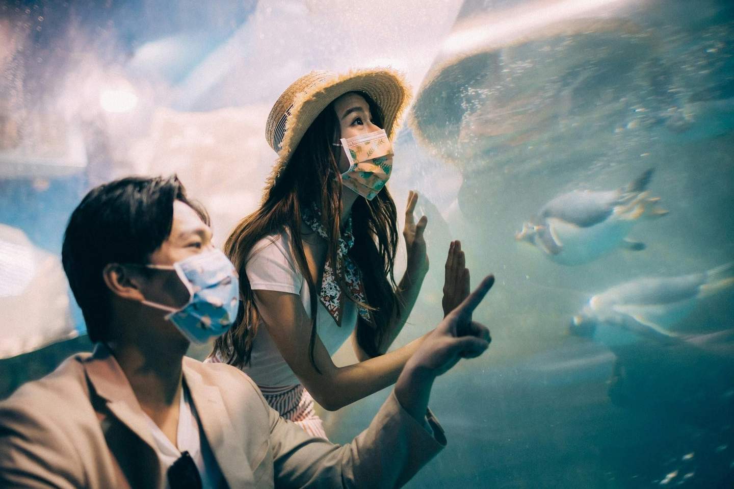 Two people wearing masks observe penguins swimming behind aquarium glass, with expressions of fascination and wonder.