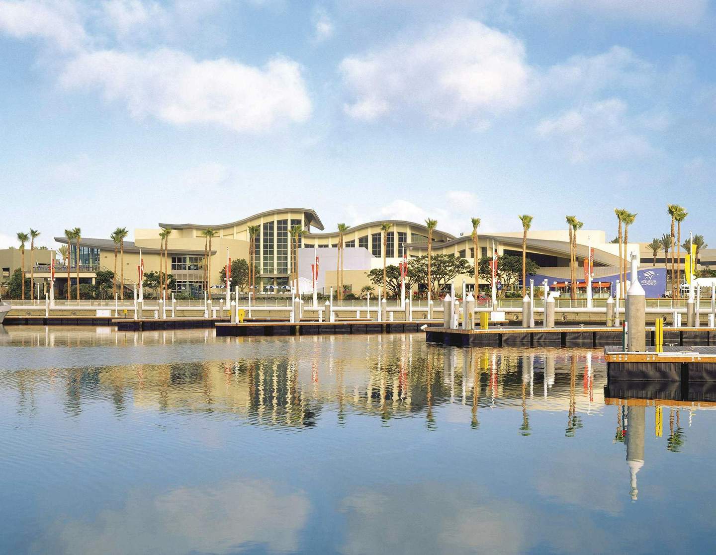 Aquarium of the Pacific A waterside view of a modern building with palm trees, docks, and calm water reflecting the scene under a partly cloudy sky.