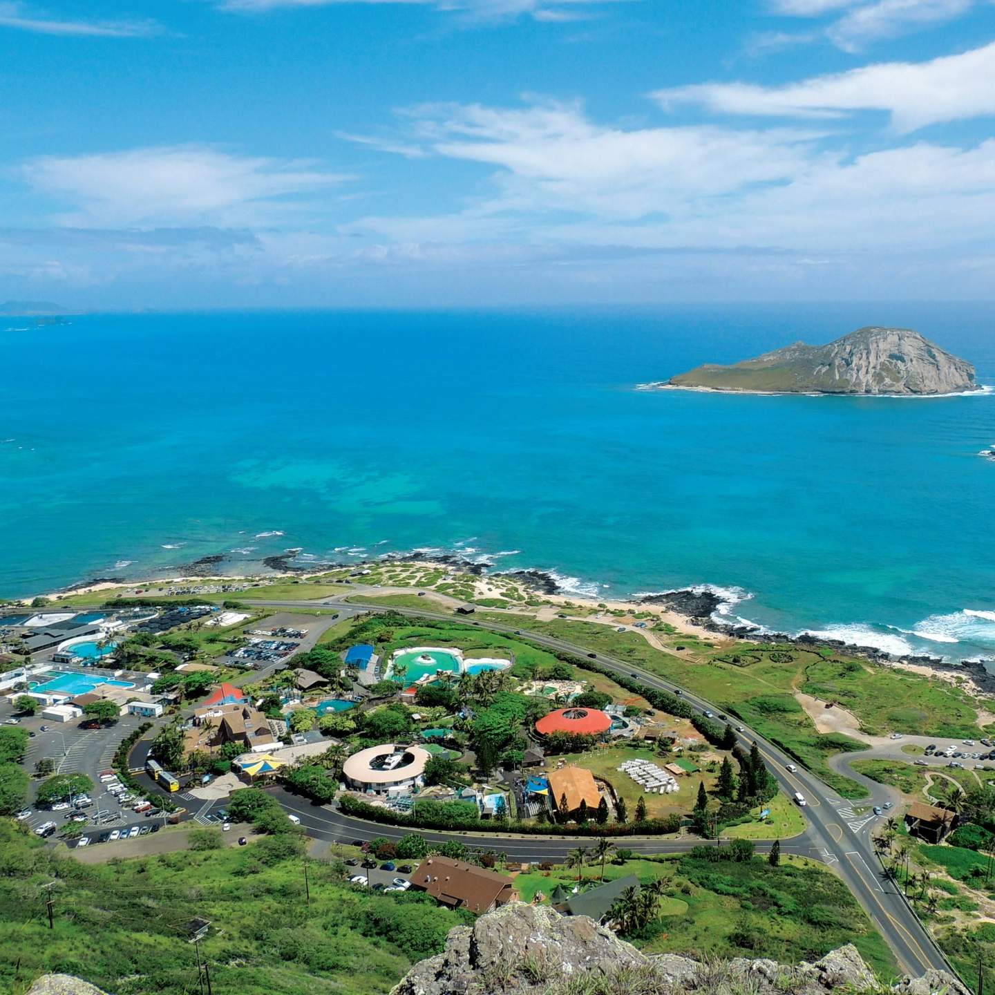 Overhead view of coastal landscape with blue ocean, small island, buildings, roads, and greenery under a partly cloudy sky.