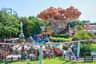 Parque de atracciones con coloridas atracciones, una gran estructura arbórea y gente disfrutando de diversas atracciones bajo un cielo azul despejado.